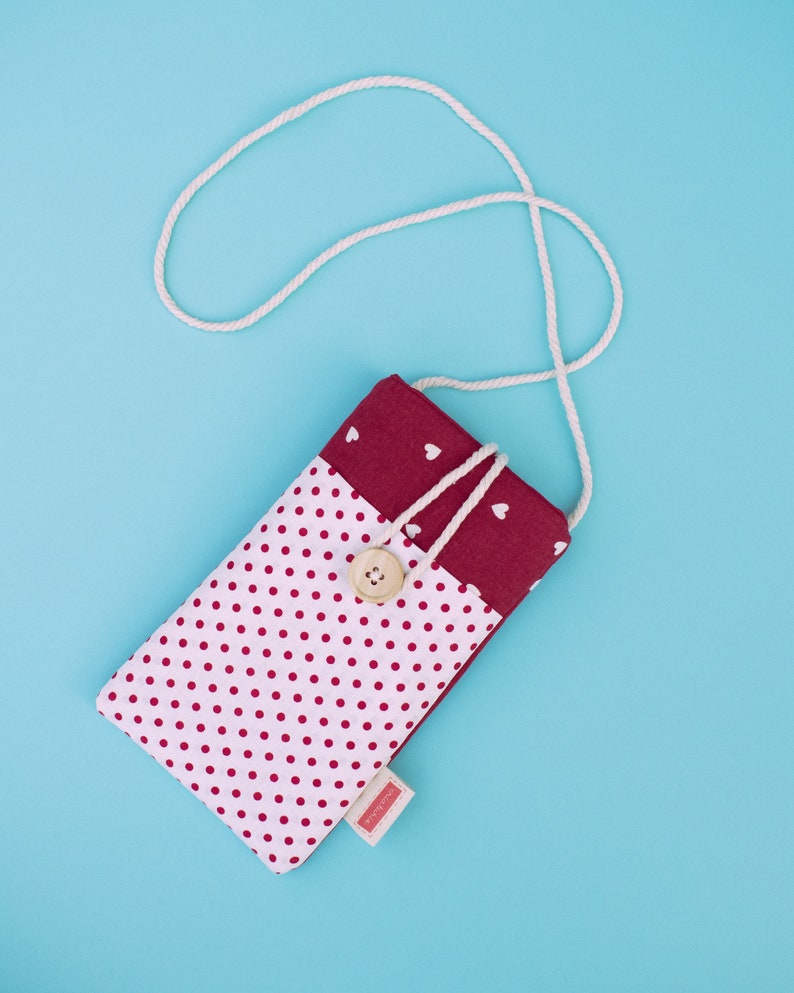 May include: A handmade, rectangular phone case with a white polka dot pattern on a light pink background. The top section is a solid burgundy color with white hearts. A wooden button secures the flap, and a white cord serves as a strap.