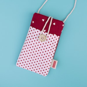 May include: A handmade phone case with a white polka dot pattern on a light pink background. The top section is a solid burgundy color with white hearts. It has a wooden button closure and a white rope strap.