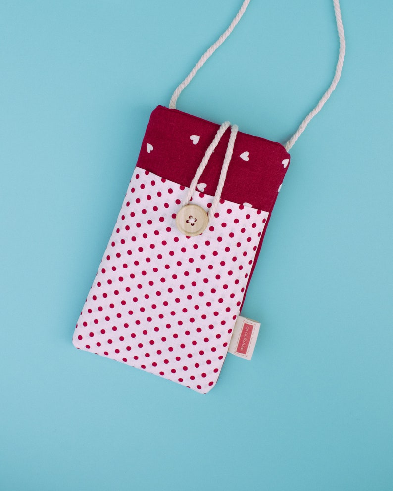 May include: A handmade phone case with a white polka dot pattern on a light pink background. The top section is a solid burgundy color with white hearts. It has a wooden button closure and a white rope strap.