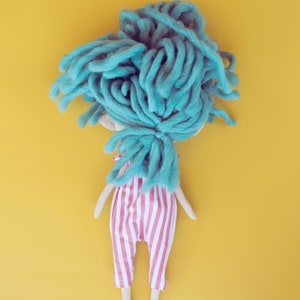 May include: A handmade doll with a light blue yarn wig and a red and white striped jumpsuit. The doll has a simple, whimsical design with a yellow background.