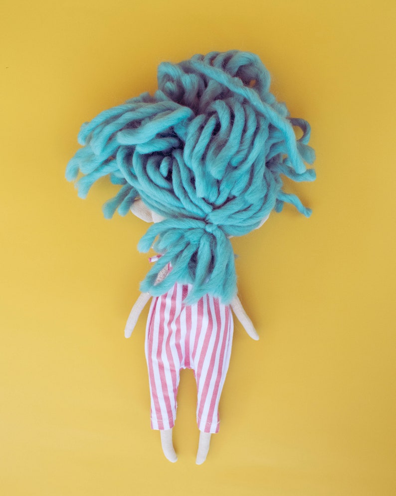 May include: A handmade doll with a light blue yarn wig and a red and white striped jumpsuit. The doll has a simple, whimsical design with a yellow background.