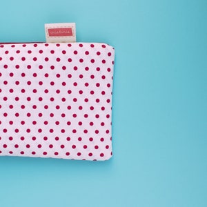 May include: A white pouch with a pattern of evenly spaced red polka dots. The pouch has a small tag with the text "criaturis" in a red rectangle. The background is a solid light blue.