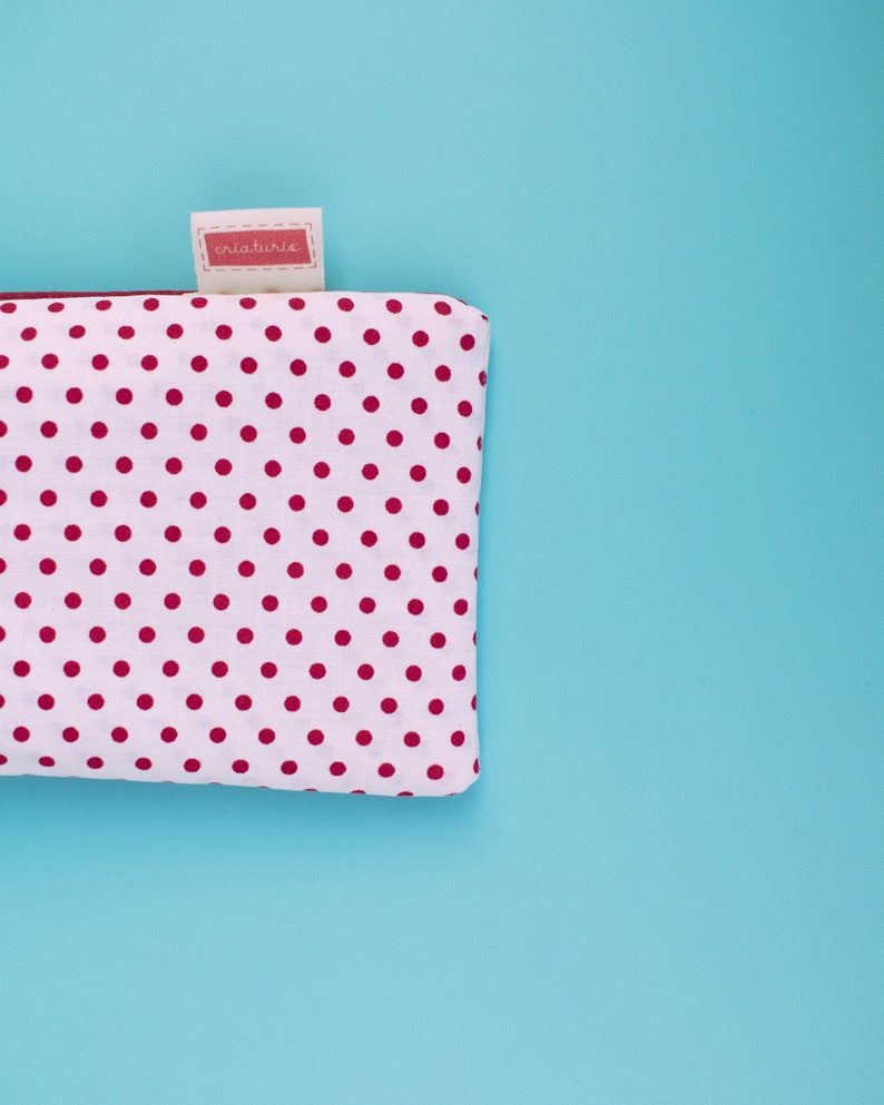 May include: A white pouch with a pattern of evenly spaced red polka dots. The pouch has a small tag with the text "criaturis" in a red rectangle. The background is a solid light blue.