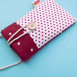 May include: A rectangular fabric phone case with a white polka dot pattern on a white background. The bottom section is a solid burgundy color with white hearts. A beige button and a white rope closure are visible. The label says "criaturita".