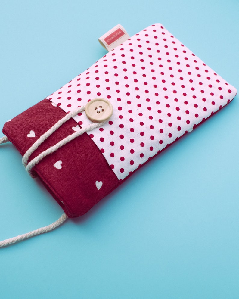 May include: A rectangular fabric phone case with a white polka dot pattern on a white background. The bottom section is a solid burgundy color with white hearts. A beige button and a white rope closure are visible. The label says "criaturita".