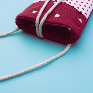 May include: A burgundy fabric phone case with a white polka dot pattern and white heart accents. The case has a white rope strap and a wooden button closure. The background is a light blue.
