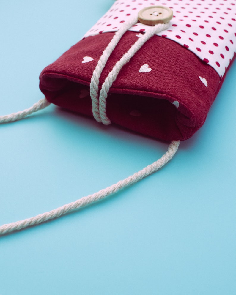 May include: A burgundy fabric phone case with a white polka dot pattern and white heart accents. The case has a white rope strap and a wooden button closure. The background is a light blue.