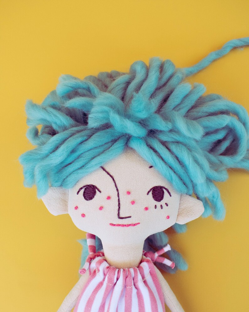 May include: A handmade doll with a whimsical design. The doll features a light-colored fabric face with embroidered details, including a simple smile and freckles. It has bright blue yarn hair and a striped pink and white dress. The background is a solid yellow.