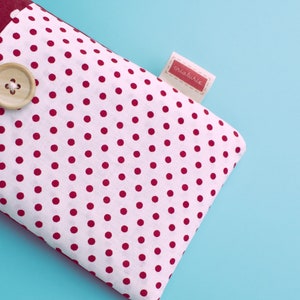 May include: A white fabric item with a red polka dot pattern, a wooden button, and a small tag with the word "criaturie" in red. The item has a burgundy border and is set against a light blue background.