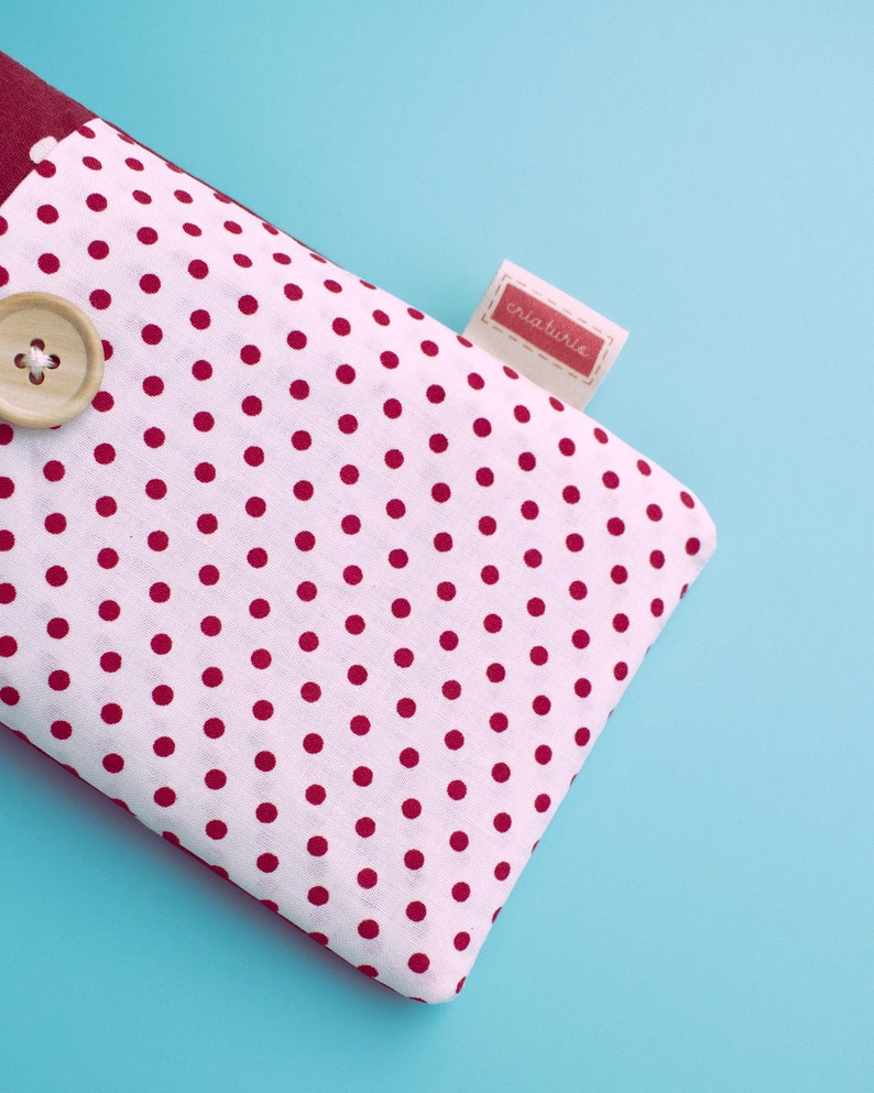 May include: A white fabric item with a red polka dot pattern, a wooden button, and a small tag with the word "criaturie" in red. The item has a burgundy border and is set against a light blue background.