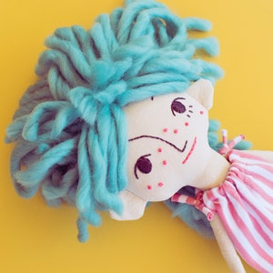 May include: A handmade doll with a light blue yarn hairstyle and a pink and white striped dress. The doll has embroidered facial features, including dark purple eyes and red freckles. The background is a solid yellow.