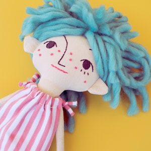 May include: A handmade doll with a light-colored face, embroidered features, and blue yarn hair. The doll wears a pink and white striped dress. The background is a solid yellow.