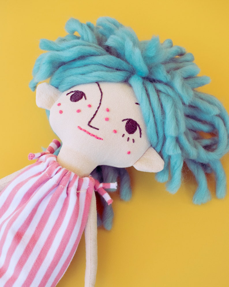 May include: A handmade doll with a light-colored face, embroidered features, and blue yarn hair. The doll wears a pink and white striped dress. The background is a solid yellow.