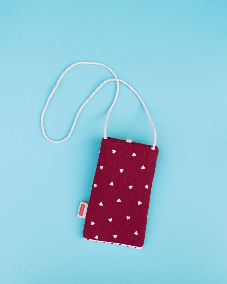 May include: A burgundy fabric phone case with a white heart pattern and a white rope strap. The case has a white dotted trim and a small rectangular tag on the side. The background is a solid light blue.