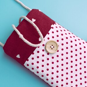 May include: A handmade fabric phone case with a white polka dot pattern on a white background. The top section is a solid burgundy color with white heart shapes. A wooden button and a white rope strap are attached.