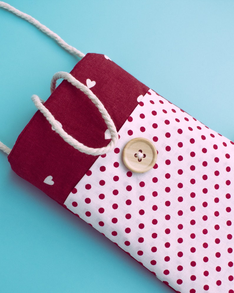 May include: A handmade fabric phone case with a white polka dot pattern on a white background. The top section is a solid burgundy color with white heart shapes. A wooden button and a white rope strap are attached.