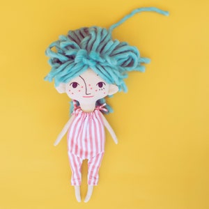 May include: A handmade doll with a whimsical design. The doll has a light skin tone, teal yarn hair, and a red and white striped jumpsuit. The doll's face features a simple, hand-drawn expression with freckles and a small smile. The background is a solid yellow.