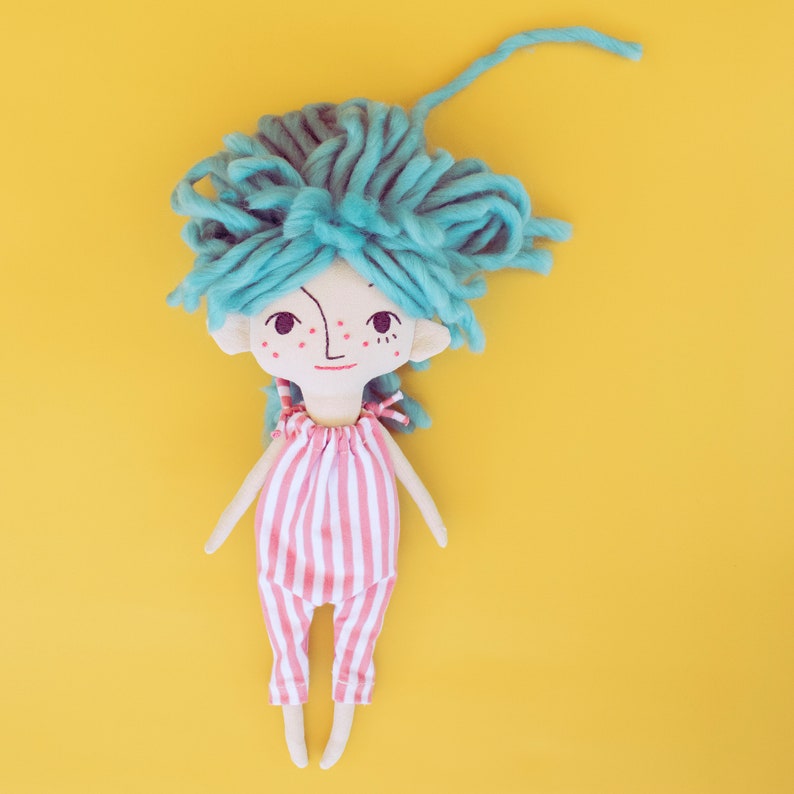 May include: A handmade doll with a whimsical design. The doll has a light skin tone, teal yarn hair, and a red and white striped jumpsuit. The doll's face features a simple, hand-drawn expression with freckles and a small smile. The background is a solid yellow.