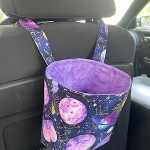 May include: A fabric car trash bag featuring a dark blue outer layer with a pattern of purple planets, stars, and gold constellations. The interior is lined with a solid purple fabric. Two fabric straps attach the bag to a car headrest.