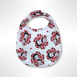 Puede incluir: Babero blanco para bebé con un estampado de corazones rojos y personajes de dibujos animados. El babero tiene forma redondeada con un agujero para el cuello. Los personajes de dibujos animados están en un círculo de lunares rojos rodeado de corazones rojos.