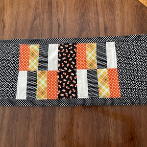 May include: A rectangular Halloween-themed table runner. The runner features a black background with white polka dots. The center has a patchwork design with orange polka dots, candy corn, and plaid patterns. White and black vertical stripes separate the patterns.