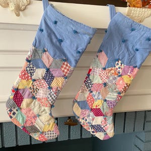 May include: Two Christmas stockings featuring a light blue fabric cuff with small blue tufted accents. The lower sections are constructed from a patchwork of various multi-colored fabric scraps in geometric shapes, joined with blue tufted knots.