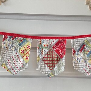 May include: A fabric bunting banner featuring seven pennants made from various patchwork quilt squares in shades of red, yellow, blue, and white, all attached to a continuous bright red ribbon for hanging across a horizontal space.