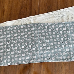 May include: Two rectangular table runners. The top runner is white with a floral pattern. The bottom runner is a light teal color with a white daisy pattern. Both are on a wooden table.