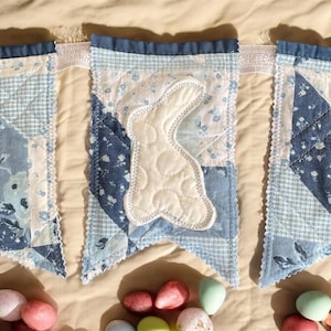 May include: A blue and white quilted Easter banner with a white bunny applique. The banner is made of various blue patterned fabrics and has a scalloped edge. Colorful Easter eggs are scattered below the banner.