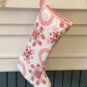 May include: A quilted Christmas stocking featuring a white background with a repeating pattern of pink floral clusters and circular leaf wreaths. The stocking has a pink fabric trim along the top opening and a matching pink fabric hanging loop.