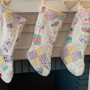 May include: Three Christmas stockings crafted from quilted fabric featuring a patchwork design of various pastel-colored floral and checkered patterns. Each stocking has a curved, arched center panel and is finished with white fabric trim.