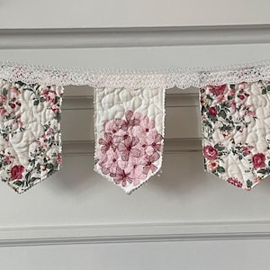 May include: A quilted fabric bunting banner featuring seven pennants suspended from a white lace trim. The pennants display alternating patterns of pink floral prints, green leafy vines, and a central cluster of pink hydrangea blossoms on white.
