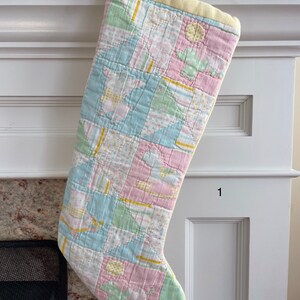 May include: A quilted Christmas stocking featuring a patchwork design in pastel shades of pink, blue, green, and yellow. The fabric displays patterns of small animals, geometric shapes, and stripes. A single yellow button secures the top cuff.