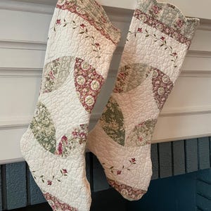 May include: Two quilted Christmas stockings featuring a cream-colored base with a patchwork design of sage green, dusty rose, and floral patterns. The fabric has a textured, stitched finish with a decorative border at the top and hanging loops.