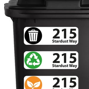 May include: A black waste bin featuring three rectangular white adhesive labels. Each label displays the address 215 Stardust Way alongside a unique icon: a black trash can, a green recycling symbol, and an orange leaf symbol for organic waste.