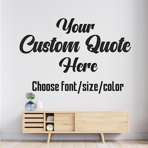 May include: A wall decal with the text "Your Custom Quote Here" in a stylish script font. Below, the text "Choose font/size/color" is displayed. The decal is shown on a white wall above a wooden cabinet.