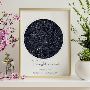 Può includere: Stampa incorniciata in oro con una mappa stellare blu scuro in un cerchio. Il testo "The night we meet" è sotto, con nomi e una data. Un elemento decorativo per la casa.