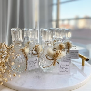 Puede incluir: Un grupo de botellas de vidrio transparente con superficies texturizadas, decoradas con flores secas y cuerda. Cada botella tiene una pequeña etiqueta blanca con el texto "Julianna Ariella Mr. Bautista 08.27.2022".