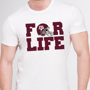 May include: White t-shirt with the maroon text "FOR LIFE" and a maroon football helmet graphic. The helmet has the letters "ATM" on the side. The shirt is a classic crew neck style.