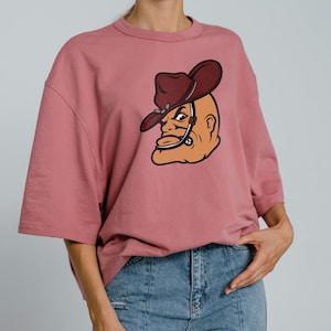 May include: A dusty rose-colored t-shirt with a large graphic of a cartoon cowboy's head. The cowboy wears a brown hat and has a stern expression. The shirt has short sleeves and a relaxed fit, suitable for casual wear.