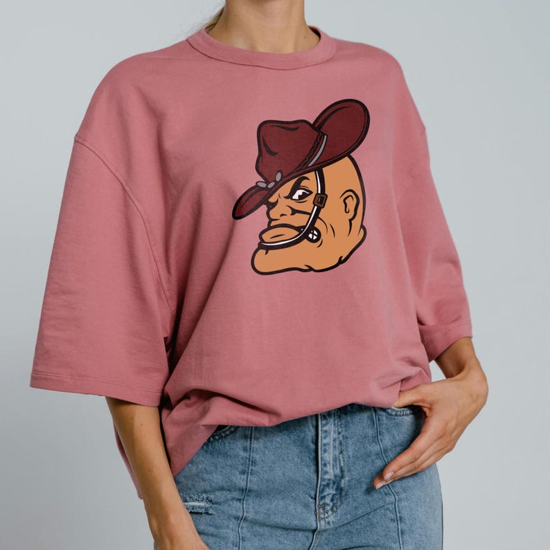 May include: A dusty rose-colored t-shirt with a large graphic of a cartoon cowboy's head. The cowboy wears a brown hat and has a stern expression. The shirt has short sleeves and a relaxed fit, suitable for casual wear.