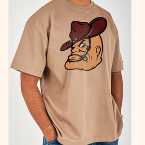 May include: A tan-colored t-shirt featuring a cartoon image of a bald man wearing a cowboy hat. The hat is dark red with a silver band. The man's face is tan with black outlines and details. The shirt has short sleeves and a crew neck.
