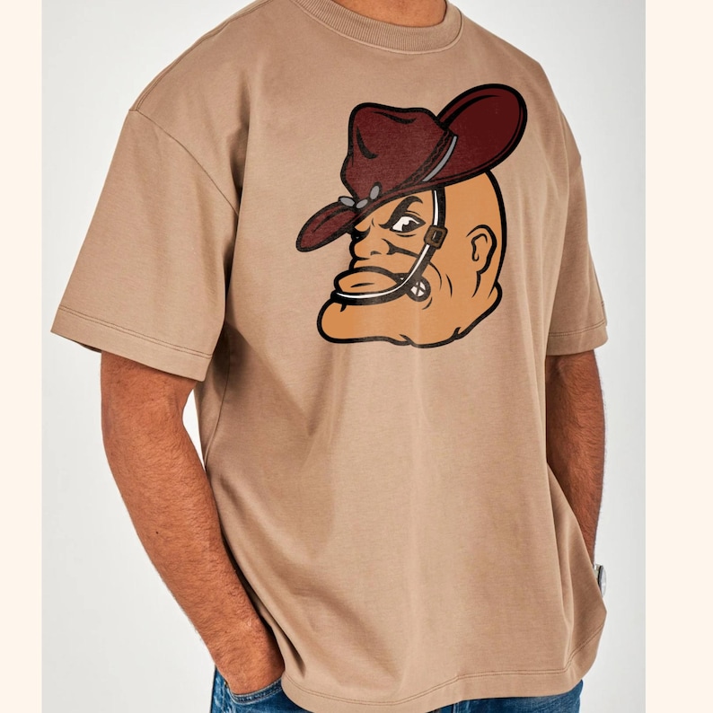 May include: A tan-colored t-shirt featuring a cartoon image of a bald man wearing a cowboy hat. The hat is dark red with a silver band. The man's face is tan with black outlines and details. The shirt has short sleeves and a crew neck.