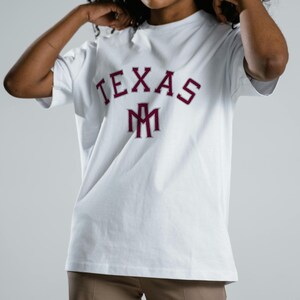 May include: White t-shirt with the word "TEXAS" in maroon block letters and a maroon logo. The shirt is a crew neck style and appears to be made of cotton. The shirt is being worn.