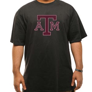 May include: Dark gray t-shirt with the letters "A", "T", and "M" in maroon. The letters are in a block style with a white outline. The shirt has short sleeves and a crew neck.