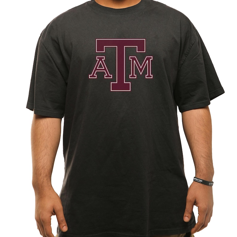 May include: Dark gray t-shirt with the letters "A", "T", and "M" in maroon. The letters are in a block style with a white outline. The shirt has short sleeves and a crew neck.