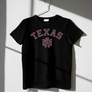 May include: Black t-shirt with the word "TEXAS" arched across the chest in maroon lettering with a white outline. Below the word "TEXAS" is a maroon and white emblem. The t-shirt is hanging on a white hanger.