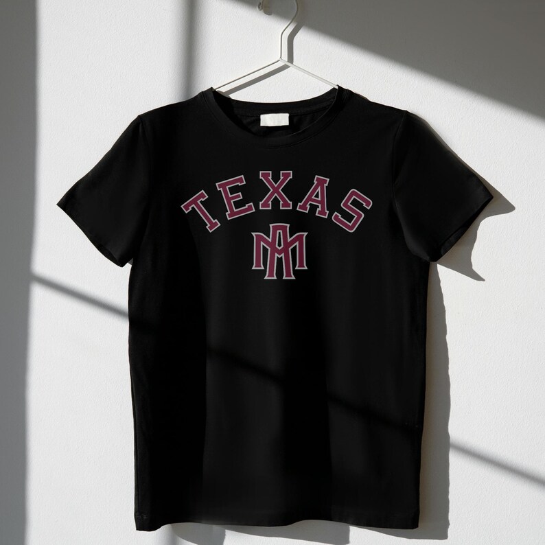 May include: Black t-shirt with the word "TEXAS" arched across the chest in maroon lettering with a white outline. Below the word "TEXAS" is a maroon and white emblem. The t-shirt is hanging on a white hanger.