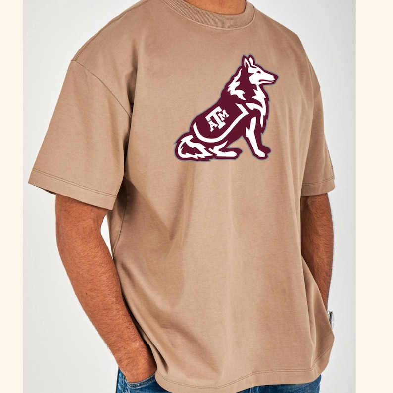 May include: A tan-colored t-shirt featuring a maroon and white graphic of a wolf with the letters "A&M" inside. The shirt has short sleeves and a crew neck.