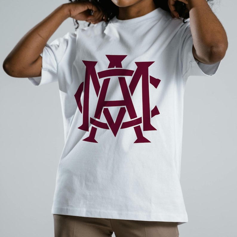 May include: White t-shirt with a maroon monogram design. The graphic features intertwined letters, creating a bold and eye-catching emblem. The shirt is made of a soft material and has short sleeves.